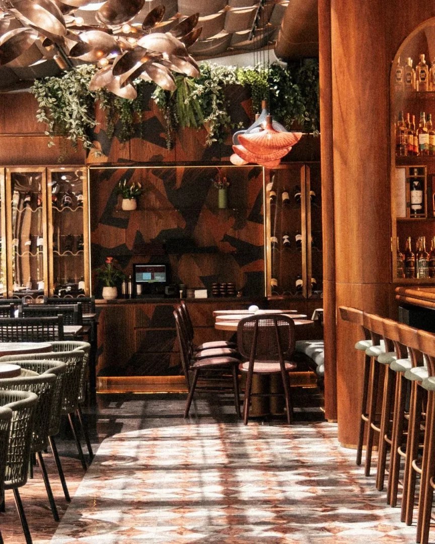 Modern ve davetkar bir atmosfer sunan bir restoran veya barın iç mekanında, şık ahşap panellerin üzerine asılmış yemyeşil bitkiler ve çarpıcı bronz avizelerle zenginleştirilmiş tavan, farklı oturma düzenlerine sahip masalar ve yüksek bar sandalyeleriyle dolu, karmaşık desenli mermer zeminde ışık yansımalarının dans ettiği sıcak ve detaylı bir tasarım.