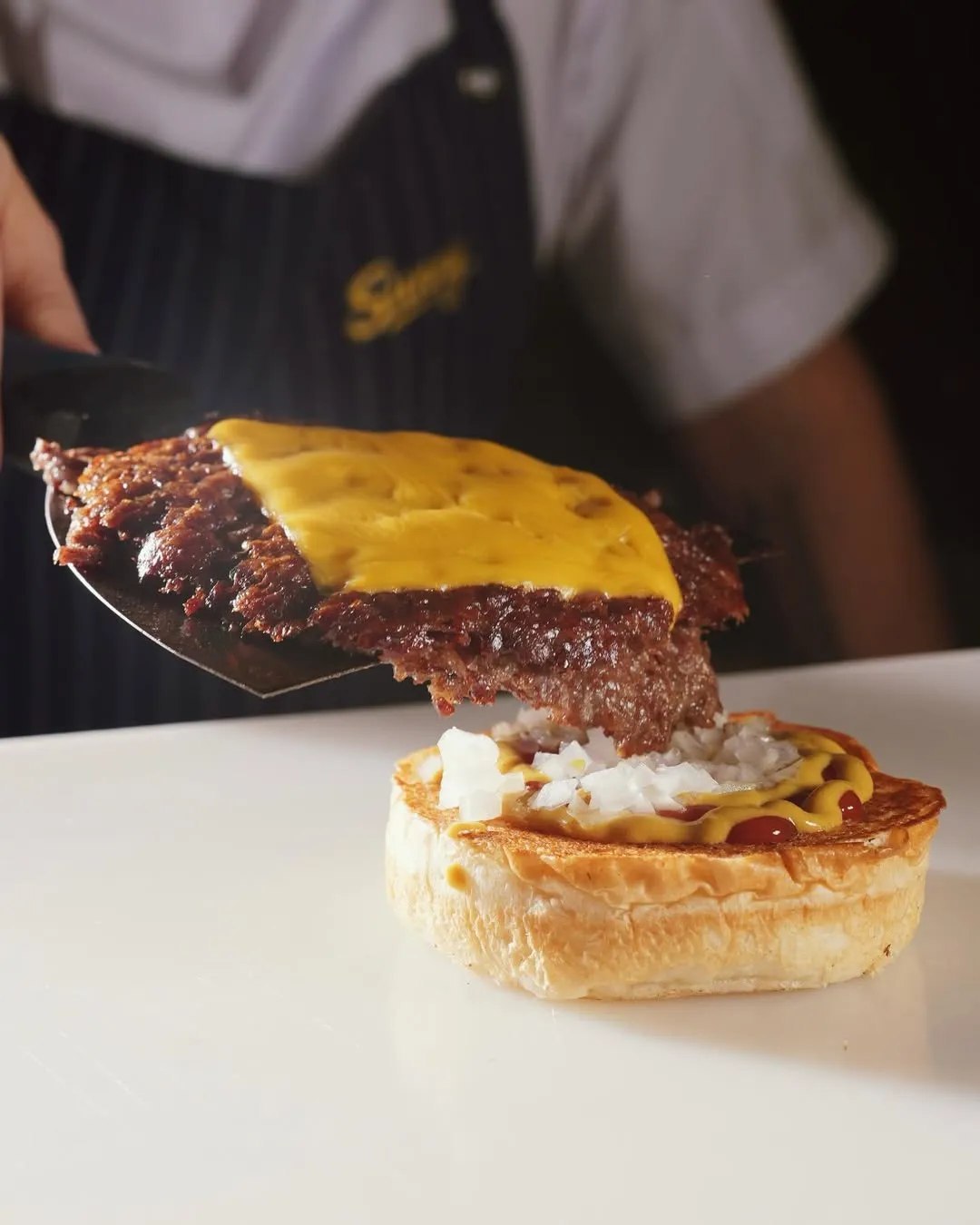Beyaz bir zeminde, erimiş peynirli, domatesli ve marullu iki köfteli, parlak ekmekli lezzetli bir smash burger, bir kesekağıdından taşan altın sarısı çıtır patates kızartmaları, küçük bir kapta kremsi sarı bir sos ve siyah pipetli şeffaf bardakta, üzerinde Sunny markası yazan ferahlatıcı limonata iştah açıcı bir şekilde duruyor.